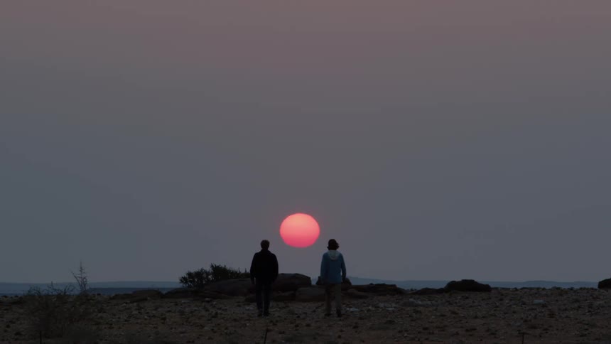 Man and woman walk across beach toward pink son floating near horizon