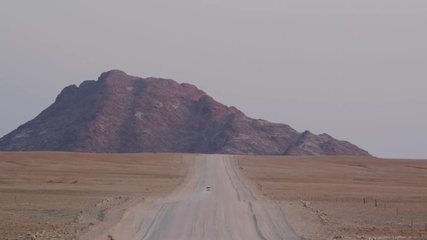 A car leaves a trail of dust as it travels on a dirt road towards a large rock formation
