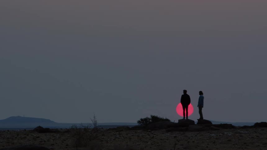 Two people stand on rocks outdoors with the sun hovering right behind them