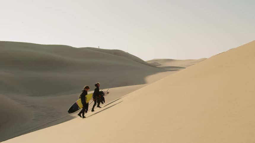 A pair of surfers in wet suits walking up a large sand dune in the desert while carrying surfboards