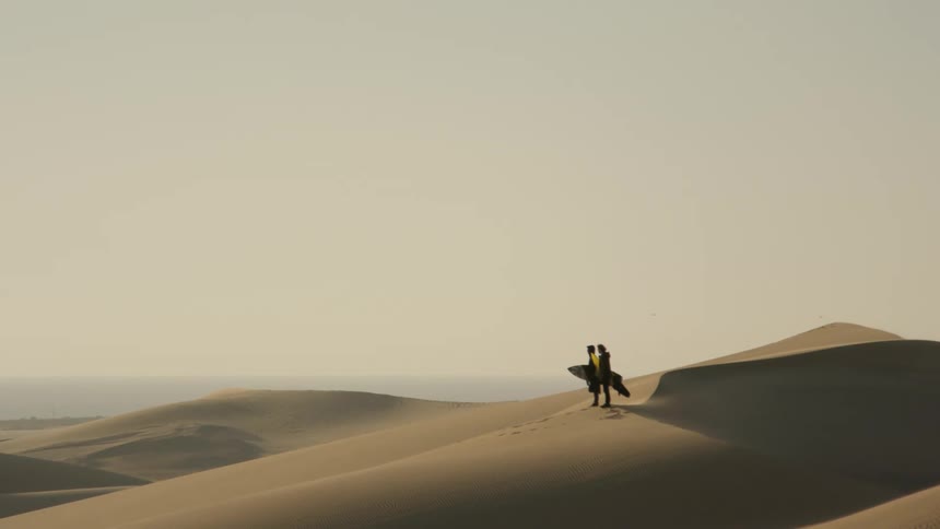 Two people standing on sand holding a surfboard with the sky in the background