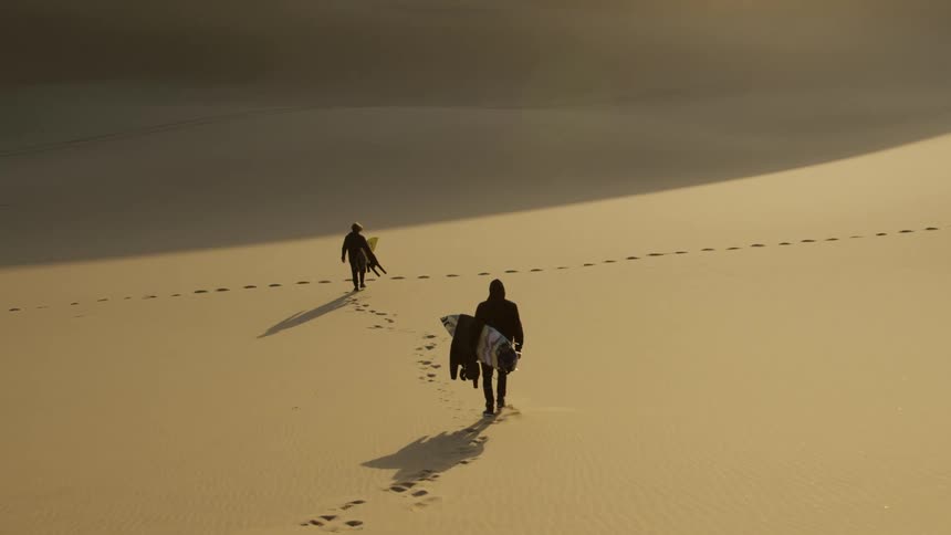 Two men holding surfboards walk downhill in the desert