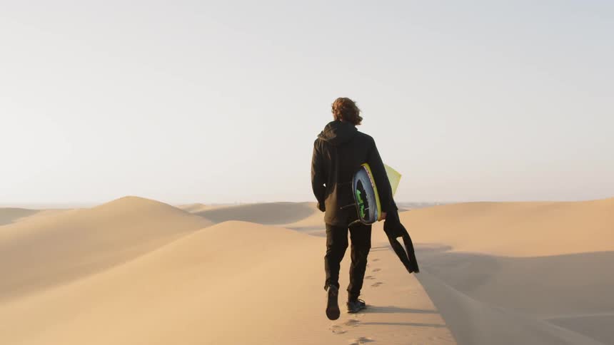 Man holding a surfboard walks on the sand dune in the desert