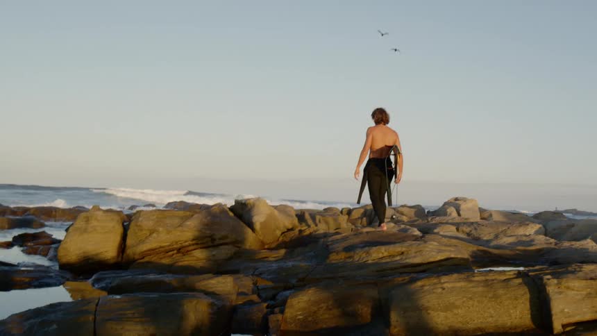 A teenager carrying his surf board walks across rocks towards the water