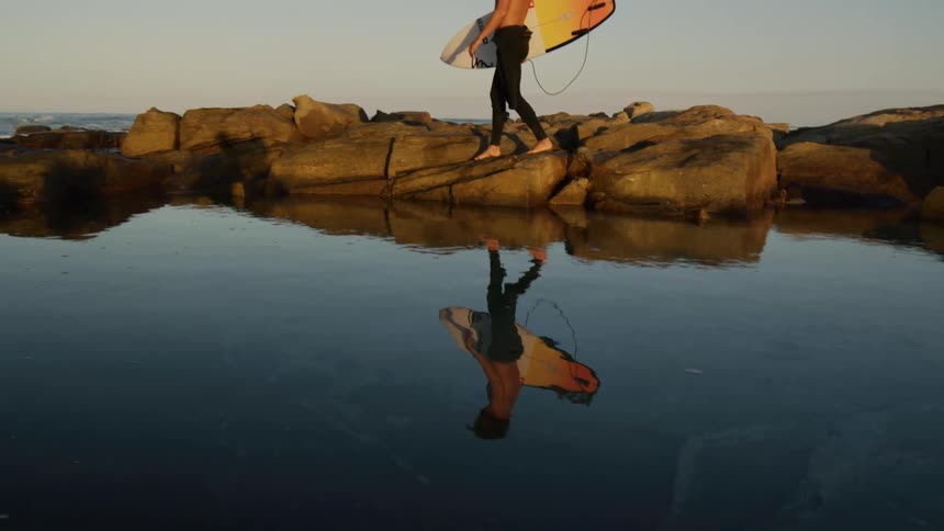 A surfer with a board walks on rocks by a pool of water