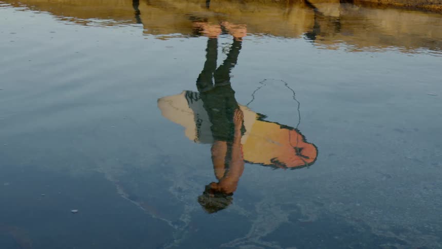 The reflection on the water of a man carrying a surfboard as he walks atop large stones