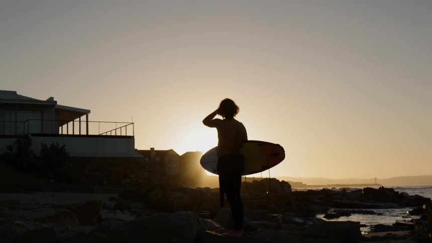 A silhouette of a man holds a surfboard and looks at the sunset