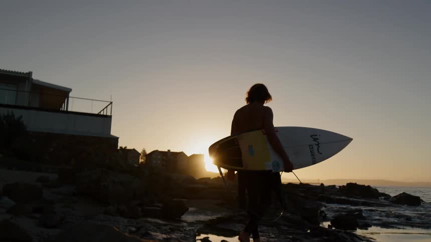 A silhouette of a male surfer holding a surfboard walks on the coast