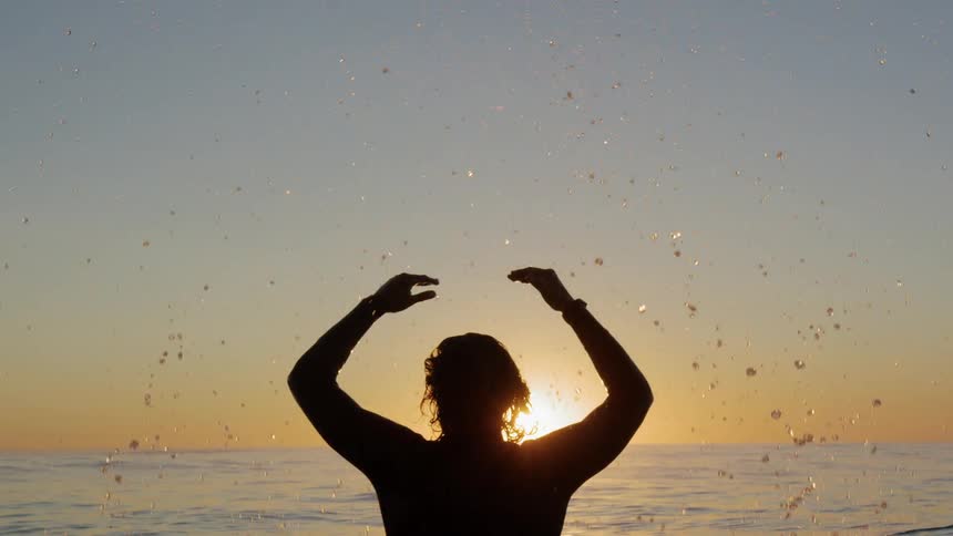 A swimmer in a wet suit standing in the ocean raises his arms splashing water at sunrise