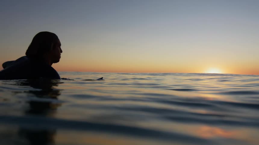 The sun sits on the horizon of the ocean as a man drifts by on the water