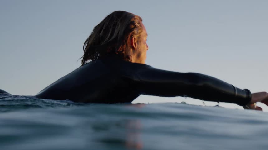 A surfer in a wet suit paddles out into the ocean on his board at sunrise