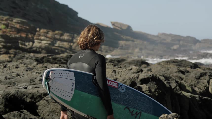 A man carries his surfboard as he walks at the bottom of a cliff near the ocean