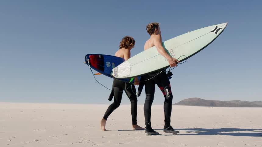 Two male surfers with their wet suits pulled up to the waist walk along the sand of the beach while carrying their surfboards