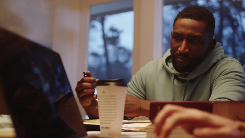 A black man sits at a table and watches a white male type on a laptop