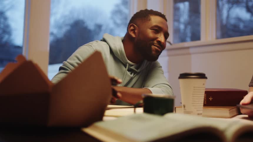 Two men sit at a table and one man shows the other something on his laptop