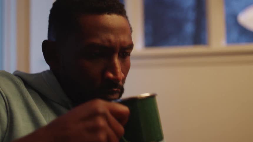 A black man takes a drink from a mug while sitting indoors