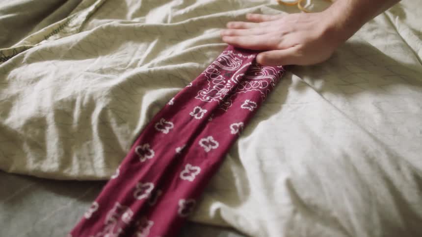 A person folding a bandana on a bed with a sheet and a pillow