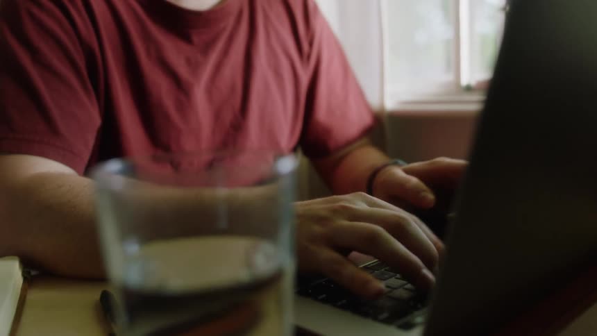 A man in a red shirt is typing on a laptop