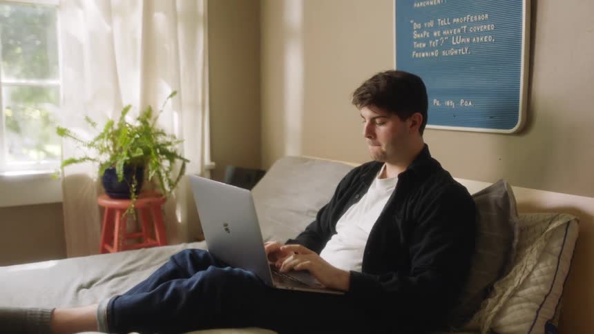 A man sits on his bed and types on his laptop