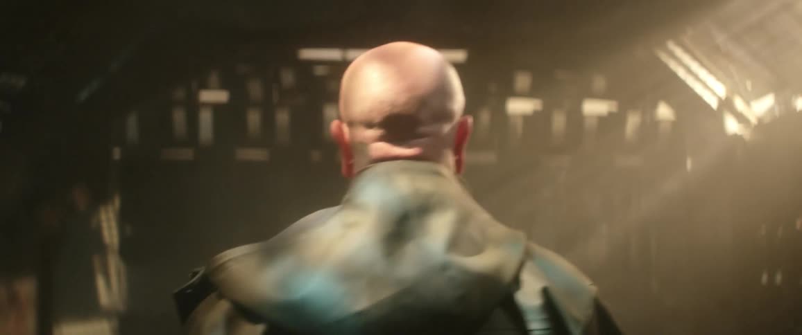 Bald man wearing a jacket with hoodie walks down a dimly lit hallway