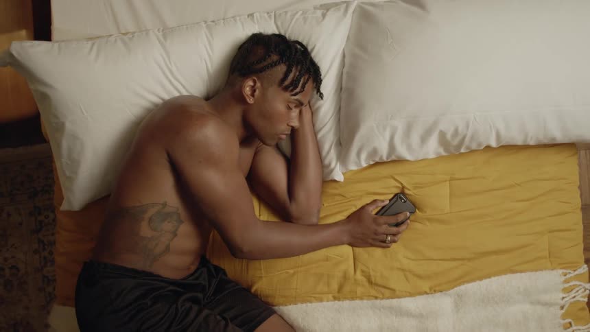 A black man sleeps on a bed while holding a smartphone