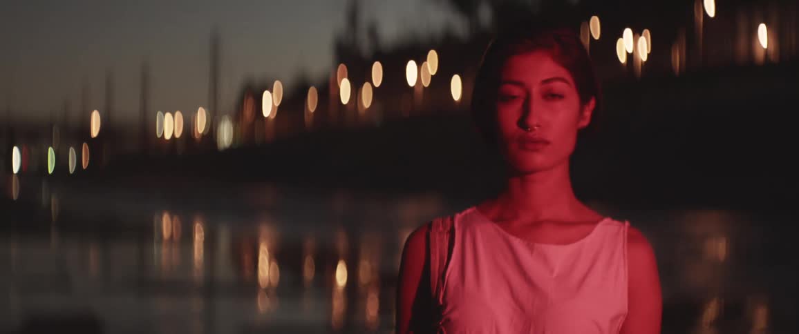 A woman standing outdoors in a city at night looking into the camera while illuminated by a flickering red light