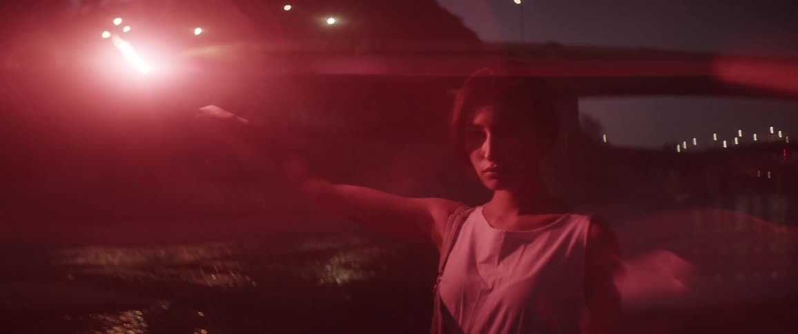 A woman holding up a lit flare with a bridge and a canal in the background
