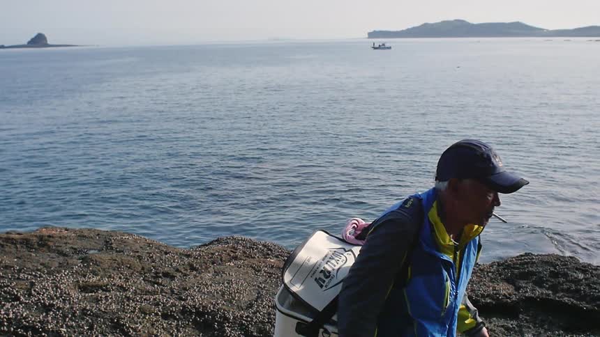 An older man smoking a cigarette and carrying a cooler walks away from a shore