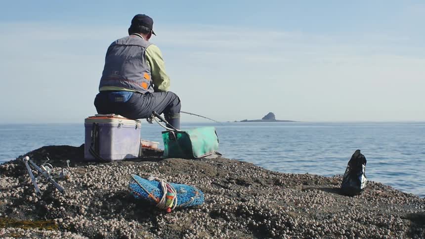 A man sitting on a cooler and fishing from a rocky shoreline