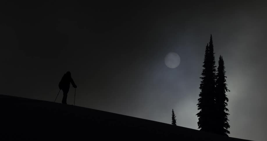 Skier stands on the top of a slope on a cloudy day with sun behind creating silhouette