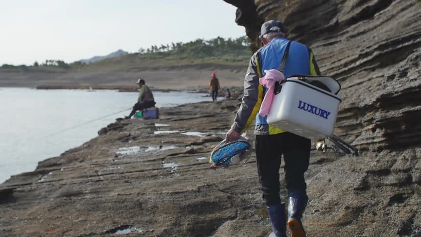An older man carries fishing gear and a cooler along a beach
