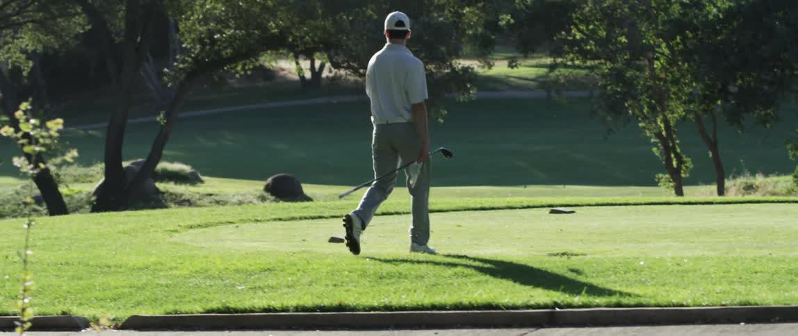A young man carrying a golf clubs strides onto a putting green