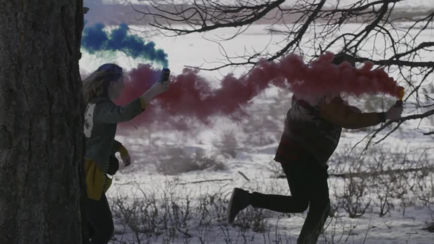 Man and woman running through the snowy wilderness while carrying colorful smoking flares