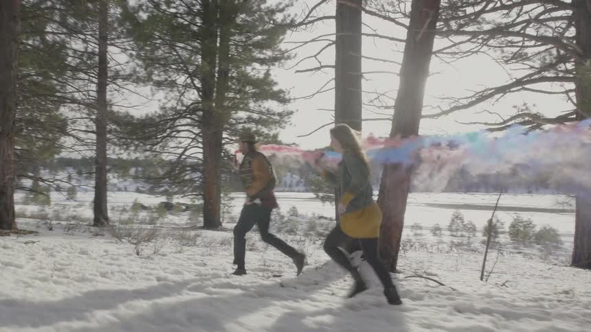 A man and woman run through a snowy and forested area with colorful smoke trailing behind them
