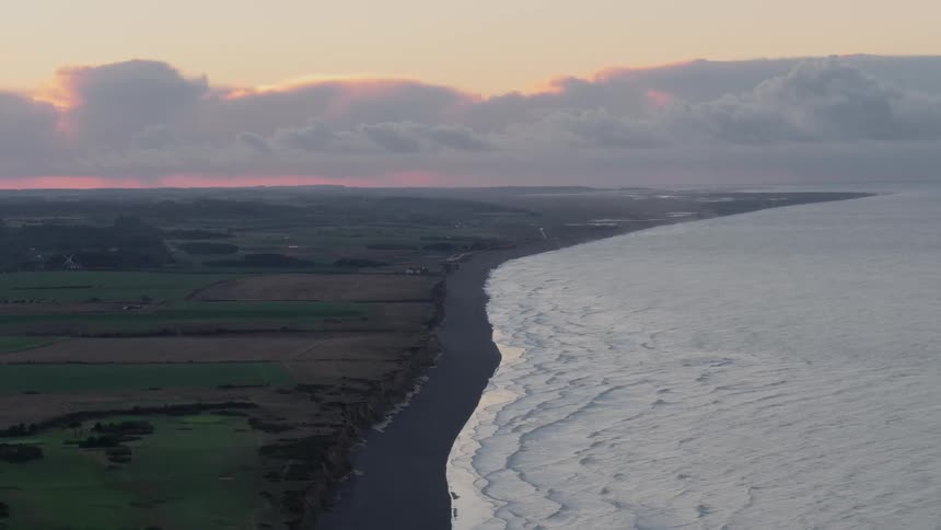 Sun turns sky behind cloud bank red as surf rolls onto dark sand beach near low cliffs