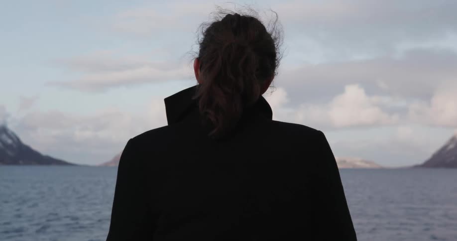 A woman in a dark coat looking out over a body of water and the distant mountains