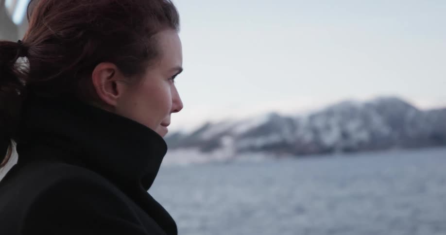 A woman with her coat collar up and hair in a ponytail looks out at the water and snow dotted land on a winter day
