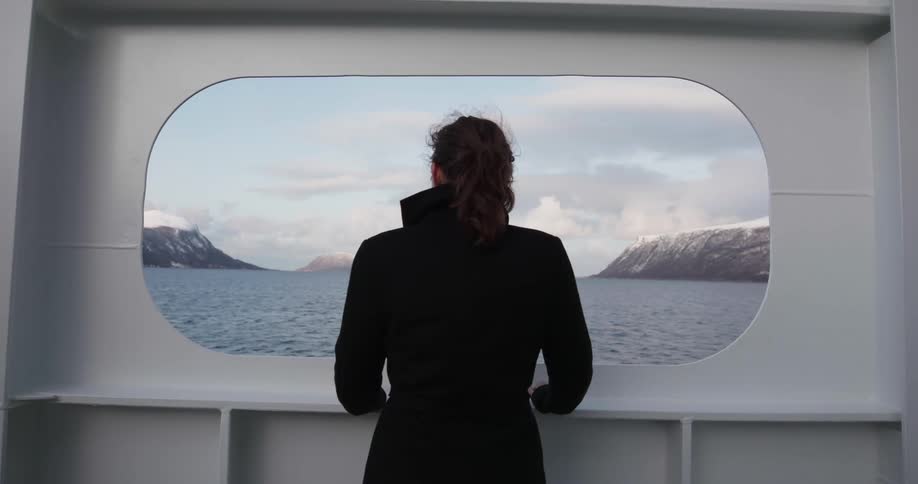 A woman looks out a window on a boat towards the snow covered mountains
