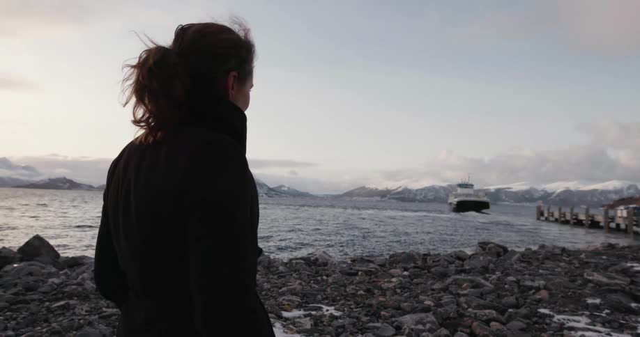 Woman stands on the pebbles beach and watches a ship traveling across the lake