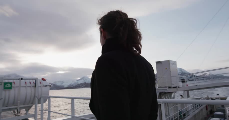 A white woman in a coat stands on a ship and looks at the mountains on the distant shore