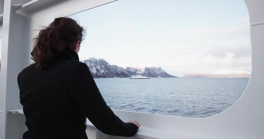 A woman stands on a boat and looks through a window at the sea and mountains