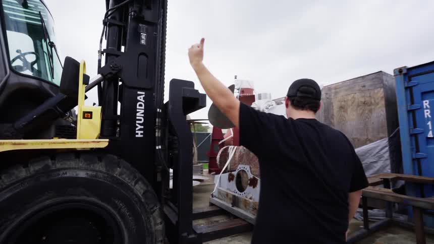 A worker helps guide a forklift driver hauling a large bladed engine