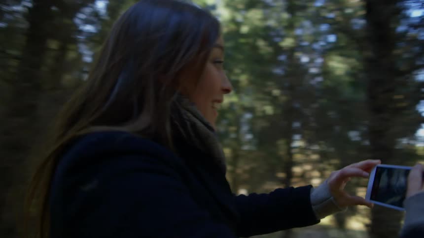 Female smiles as she holds a cell phone and tries to take a video of male while they are running through the trees of a wooded area