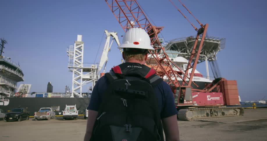A man with a hard hat and backpack walks to a shipyard