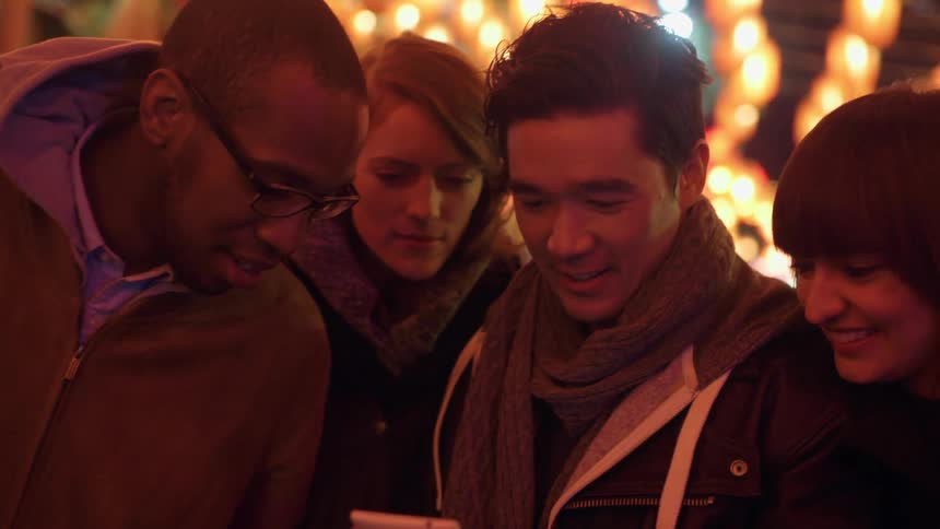A man holds his cell phone and him and his three friends look at it