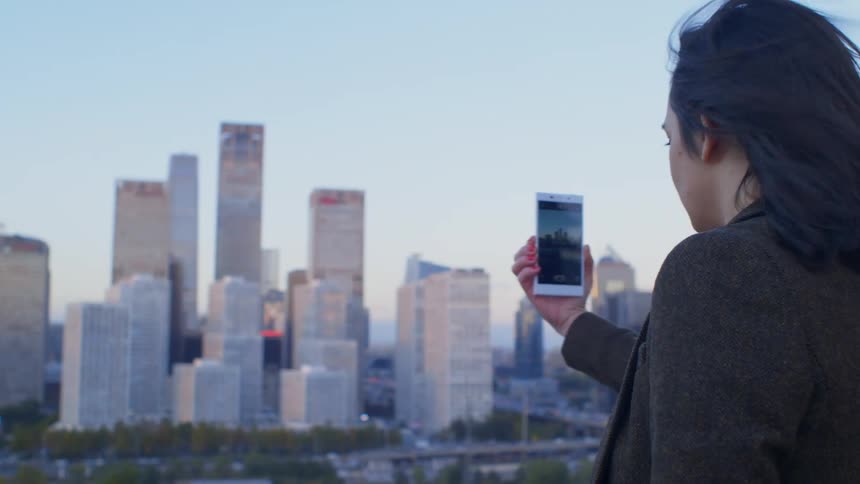 A woman holds her phone up and captures the Beijing skyline while the wind blows her hair
