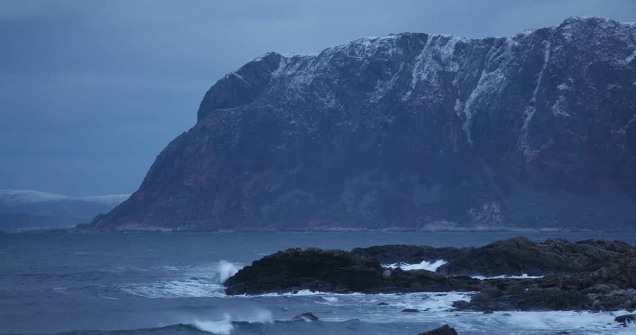 Waves crash on a rocky coast as a mountain looms high over the terrain