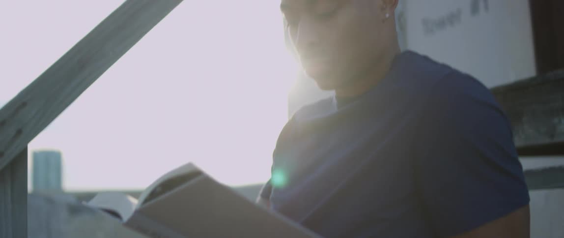 Black man sits on an outdoor step as he reads a book