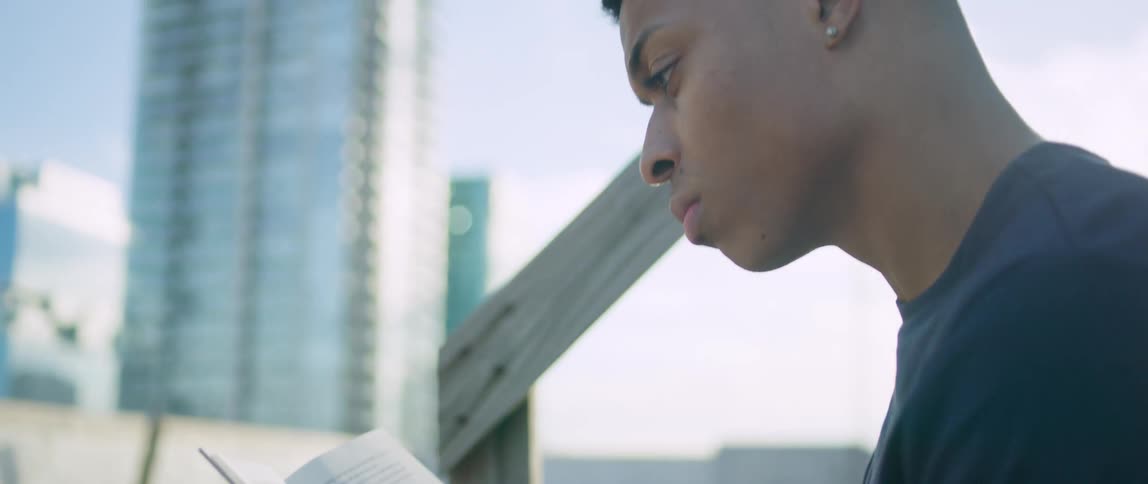 A teen sits outside and rads with skyscrapers in the background