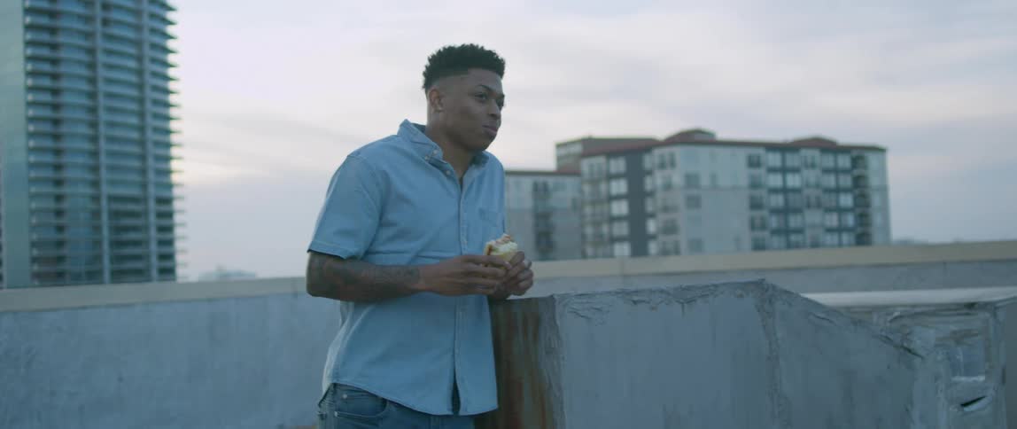 A male teenager eats a sandwich on a rooftop with skyscrapers in the background
